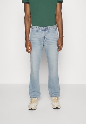 Homme portant un jean bleu clair droit, une chemise verte à manches courtes et des baskets beiges, debout devant un fond blanc uni.
