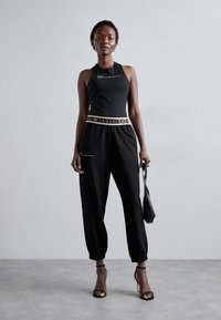 Melnā bezpiešu topiņa ar logo, apvienota ar melniem sporta biksēm ar zelta akcentu jostu. Modelis tur mazu melnu somiņu.