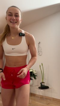 Urheilija valkoisessa Nike-urheiluliiveissä ja punaisissa shortseissa, mikrofonilla olkapäällä, hymyilee sisätiloissa kasvien ja piirroksen taustalla.