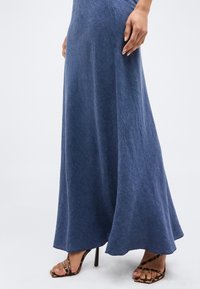 Γυναίκα που φοράει μακριά μπλε φούστα συνδυασμένη με ψηλοτάκουνα σανδάλια με λεοπάρ σχέδιο, στέκεται μπροστά σε απλό φόντο.