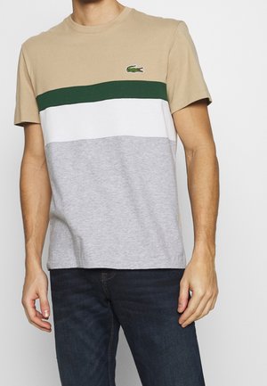 Man draagt een colorblock T-shirt met beige, groene, witte en grijze strepen, gecombineerd met donkerblauwe spijkerbroek, staand tegen een effen achtergrond.