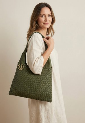 Vrouw in licht linnen jurk met een groene geweven handtas met een gouden cirkelvormig logo, iets zijwaarts gericht tegen een effen beige achtergrond.