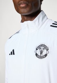 White athletic jacket featuring a zip closure, black Adidas stripes on the shoulders, and a Manchester United embroidered logo on the chest.