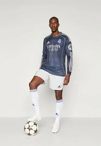 Szürke, hosszú ujjú focimez fehér részletekkel, az "Emirates" logóval, fehér rövidnadrággal és focicipőkkel, egy fekete-fehér focilabdán állva.