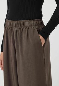 Bruine, wijde broek met een elastische tailleband en zijzakken. De stof heeft een subtiele textuur en glans. Gecombineerd met een zwart geribbeld topje.