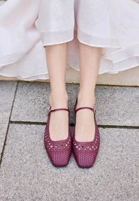 Balerini împletiți bordo cu vârful pătrat și curea pentru gleznă, având detalii decupate și o suprafață texturată, așezați pe un trotuar din piatră.