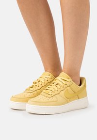 Jalat yang mengenakan sepatu Nike Air Force 1 kuning muda dengan sol putih, berdiri di atas permukaan putih.