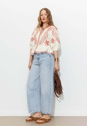 Femme debout portant un chemisier blanc avec broderie rouge, un jean large bleu clair, des sandales marron, un bracelet manchette doré et tenant un sac à franges marron.