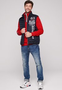 Gilet imbottito blu con testo bianco, felpa rossa sotto, jeans blu e sneakers bianche con accenti rossi. Progettato per un abbigliamento informale.