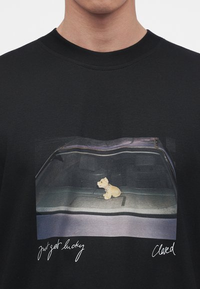 Petite figurine de chien beige assise sur le tableau de bord d'une voiture avec un pare-brise réfléchissant, sur un t-shirt noir avec les textes "just get lucky" et "Claved" en dessous.
