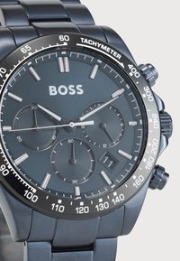 Μαύρο ρολόι από ανοξείδωτο ατσάλι με σκούρο γκρι καντράν. Περιέχει υποκαντράν χρονογράφου, κλίμακα ταχύτητας και ένδειξη ημερομηνίας στις 6 η ώρα.