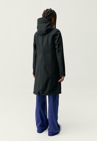 Črn plašč s kapuco z dolgim, prirejenim krojem, ki ima stranske šive in rahlo razporko na robu. Usklajen z modrimi širokimi hlačami.