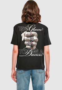 Black cotton t-shirt featuring a graphic of a hand with rings. Text reads "TOO Glam" and "TOO GIVE A Damn" in white cursive lettering.