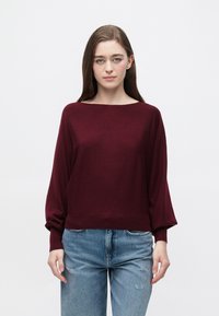 Bordowy dzianinowy sweter z szerokim dekoltem, długimi rękawami i ściągaczami, zestawiony z podniszczonymi jasnoniebieskimi jeansami.