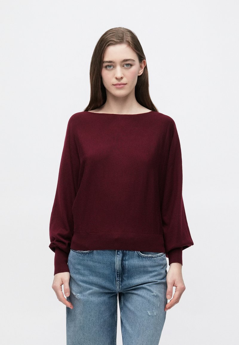 Bordowy dzianinowy sweter z szerokim dekoltem, długimi rękawami i ściągaczami, zestawiony z podniszczonymi jasnoniebieskimi jeansami.