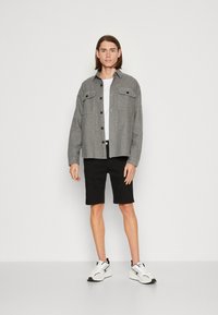Chaqueta gris de botones con dos bolsillos en el pecho, superpuesta a una camiseta blanca, combinada con pantalones cortos negros y zapatillas blancas. Fondo neutro.