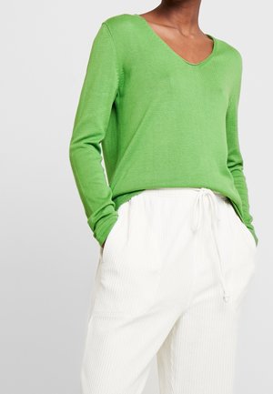 Persona que lleva un suéter de manga larga verde brillante con cuello en V y pantalones blancos con cordón, con las manos en los bolsillos, contra un fondo liso.