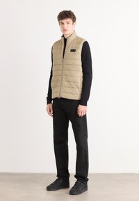 Μπεζ πλισέ γιλέκο με όρθιο γιακά και κλείσιμο με φερμουάρ, συνδυασμένο με μαύρη μπλούζα μακρύ μανίκι και παντελόνι, ολοκληρωμένο με μαύρα αθλητικά παπούτσια.