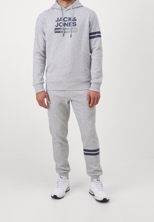 Mężczyzna ubrany w jasnoszarą bluzę z kapturem i spodnie dresowe z ciemnoniebieskimi paskami oraz napisem "JACK & JONES PERFORMANCE", do tego białe buty Puma.