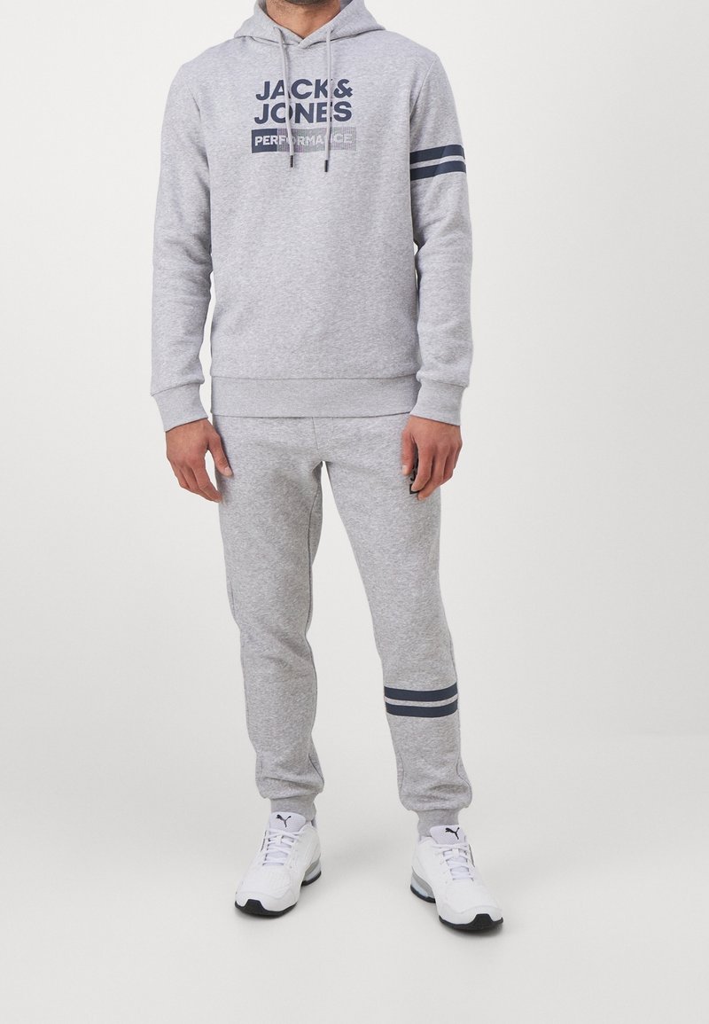 Homme portant un sweat à capuche gris clair et un jogging avec des rayures bleu foncé et le texte "JACK & JONES PERFORMANCE", associé à des baskets blanches Puma.