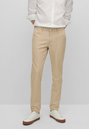 Mężczyzna ubrany w beżowe chinosy, białą koszulę zapinaną na guziki z podwiniętymi rękawami oraz białe trampki z brązowymi podeszwami stoi na tle szarego tła.