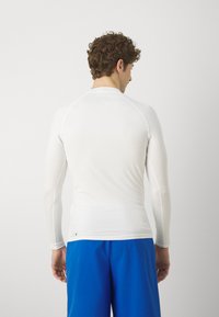 Biała, długorękawowa odzież sportowa z gładkiego, elastycznego materiału. Posiada rękawy raglanowe i dopasowany krój, zestawiona z niebieskimi szortami.