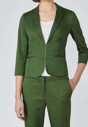 Woman wearing a fitted olive green blazer with three-quarter sleeves and matching high-waisted trousers, paired with a white top.