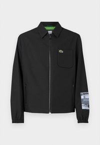Black jacket made of smooth fabric with a green inner lining, front zipper, left chest pocket, and a patterned sleeve detail.