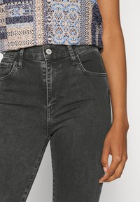 Primer plano de una persona que lleva jeans negros de talle alto y una blusa azul con un estampado, con un brazo relajado a un lado.