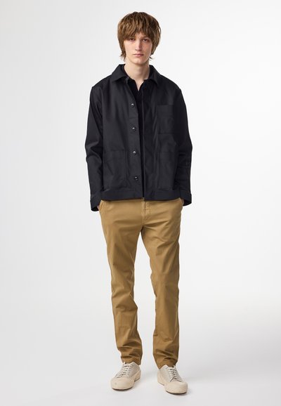 Svart lättviktsjacka med knappstängning och två framfickor, tillsammans med khakifärgade byxor och beige sneakers. Slät textur, avslappnad passform.