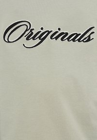 Μαύρο κείμενο σε καλλιγραφική γραφή "Originals" με λευκό περίγραμμα, κεντημένο σε ανοιχτό γκρι ύφασμα.
