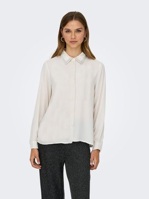 Vrouw die frontaal staat, draagt een crèmekleurige blouse met lange mouwen en knopen en donkere, getextureerde broek tegen een effen achtergrond.