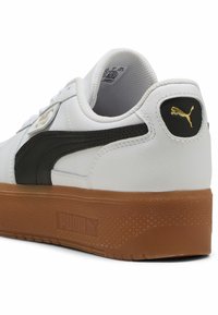 Białe sneakersy z czarnym paskiem PUMA, teksturowana brązowa gumowa podeszwa i złote akcenty logo na pięcie. Górna część z gładkiej skóry i wyściełany kołnierz.
