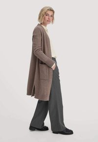Lungo cardigan marrone in morbido materiale, caratterizzato da tasche frontali e polsini a costine, indossato sopra una maglia chiara e pantaloni grigi a righe.