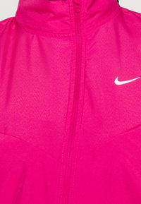 Jasnoróżowa kurtka Nike z wysokim kołnierzem, pełnym zamkiem i małym białym logo. Materiał ma gładką, lekko błyszczącą fakturę.