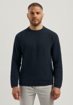 Marineblauw gebreid sweater met een textuurpatroon, ronde hals, en lange mouwen, gecombineerd met lichtgekleurde broeken.