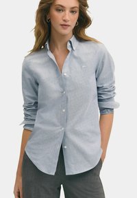 Světle modrá košile na knoflíky v jemném materiálu, s dlouhými rukávy, klasickým límcem a jemnými detaily šití. Spárována se šedými kalhotami.