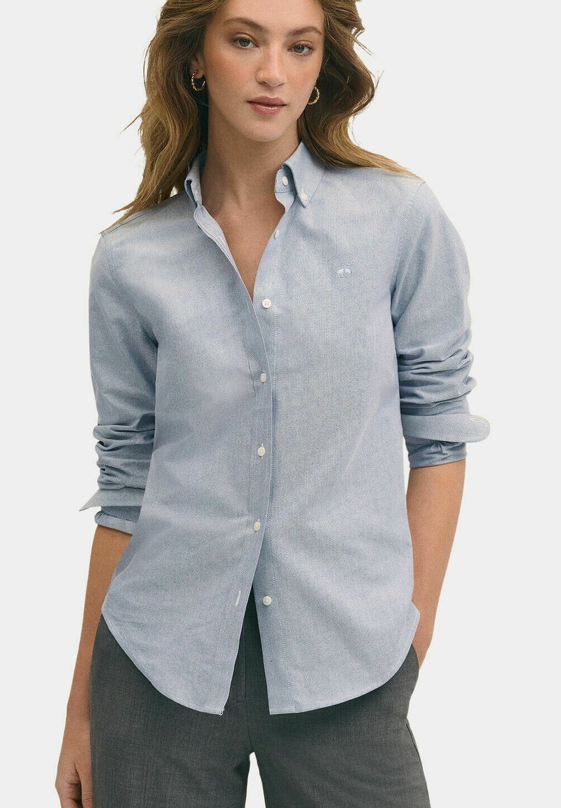 Světle modrá košile na knoflíky v jemném materiálu, s dlouhými rukávy, klasickým límcem a jemnými detaily šití. Spárována se šedými kalhotami.