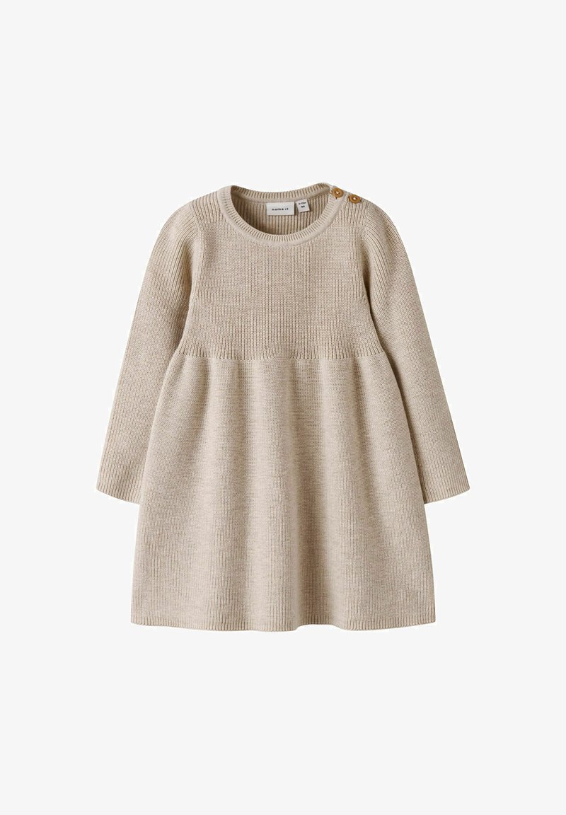 Beige kötött ruha hosszú ujjakkal, bordázott textúrával és bővülő szoknyával. Két fából készült gomb található a dekoltázsán.