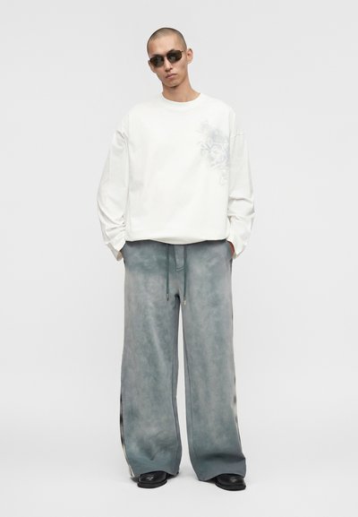 Άνδρας που φοράει λευκό πουκάμισο με μακριά μανίκια σε υπερμεγέθη μέγεθος με διακριτικό σχέδιο, άνετο γκρι παντελόνι, μαύρα παπούτσια και σκούρα γυαλιά ηλίου, στέκεται με τα χέρια στις τσέπες.