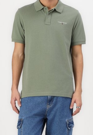 Mężczyzna ubrany w oliwkową koszulkę polo Tommy Jeans i niebieskie spodnie typu cargo z denimu, stoi z ramionami swobodnie opuszczonymi wzdłuż ciała.