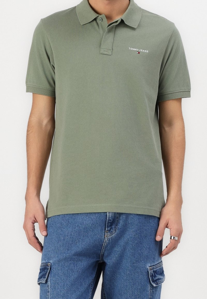 Homme portant un polo Tommy Jeans vert olive et un pantalon cargo en denim bleu, debout, les bras détendus le long du corps.