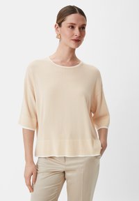 Jasny brzoskwiniowy sweter z okrągłym dekoltem, rękawami o długości trzech czwarci i białymi wykończeniami. Miękka faktura o luźnym kroju, zestawiony z beżowymi spodniami.