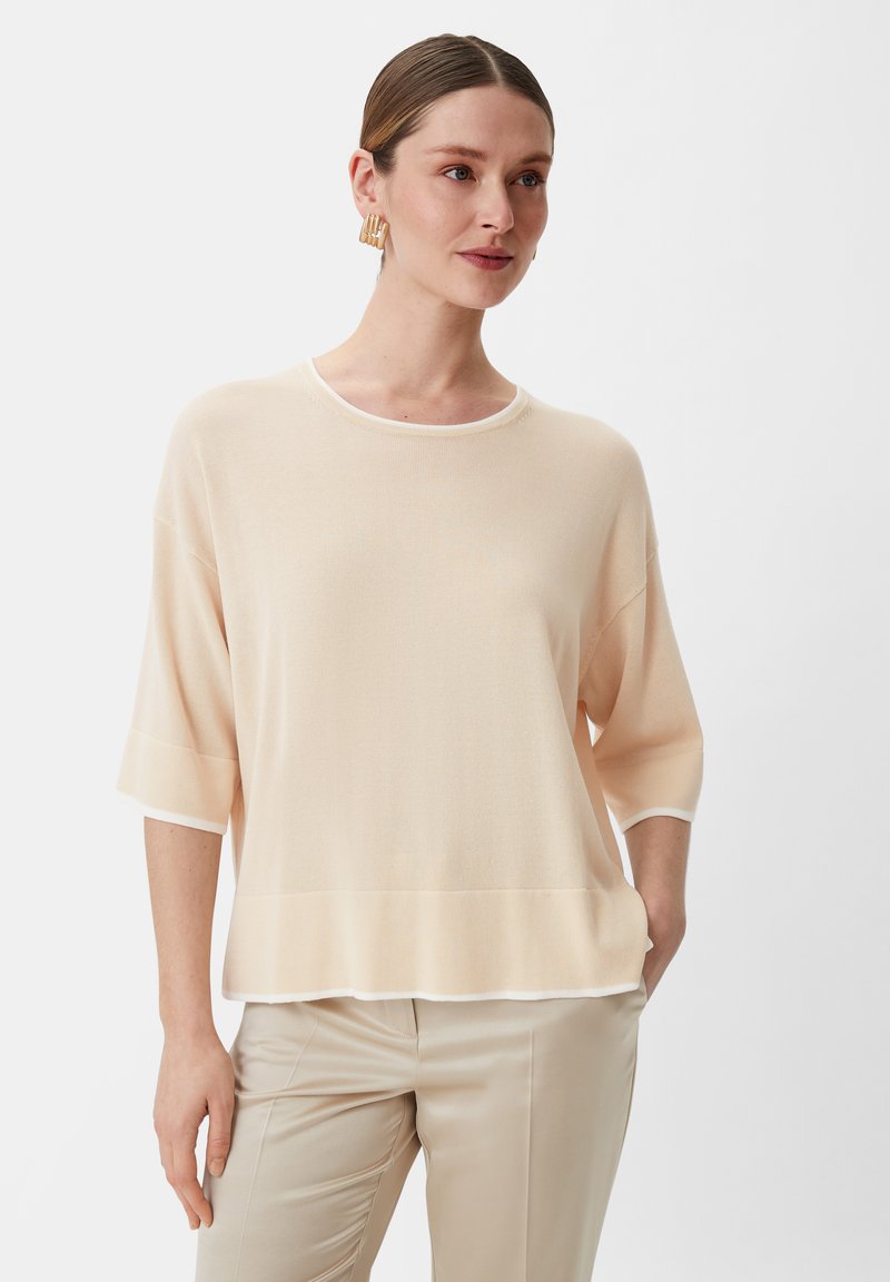 Jasny brzoskwiniowy sweter z okrągłym dekoltem, rękawami o długości trzech czwarci i białymi wykończeniami. Miękka faktura o luźnym kroju, zestawiony z beżowymi spodniami.