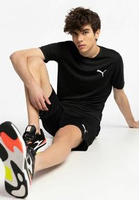 Sort atletisk skjorte og shorts med hvidt logo. Model iført sorte og multikolorede sko, siddende stilling, glat stoftekstur.