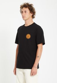 Sort t-shirt lavet af bomuld. Har et rundt orange grafisk motiv på brystet. Korte ærmer, rund hals, afslappet pasform.