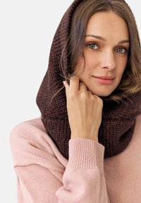 Brązowy dziany komin wokół szyi, z teksturowanym wzorem. Modelka ma na sobie miękki różowy sweter z prążkowanymi mankietami, na tle w neutralnych kolorach.