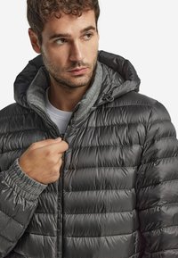 Dunkelgraue Pufferjacke mit gestepptem Design. Verfügt über eine Kapuze, graue Innenauskleidung und gerippte Bündchen für zusätzlichen Wärme- und Tragekomfort.