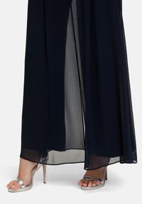 Granatowa sukienka z szyfonu z przezroczystą narzutą i wysokim rozcięciem. Metaliczne srebrne sandały na obcasie z szerokim paskiem i otwartym noskiem.