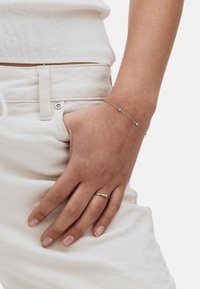 Srebrna bransoletka z małymi okrągłymi akcentami, noszona na dłoni spoczywającej na jasnym materiale; minimalistyczny design w połączeniu ze srebrnym pierścionkiem.